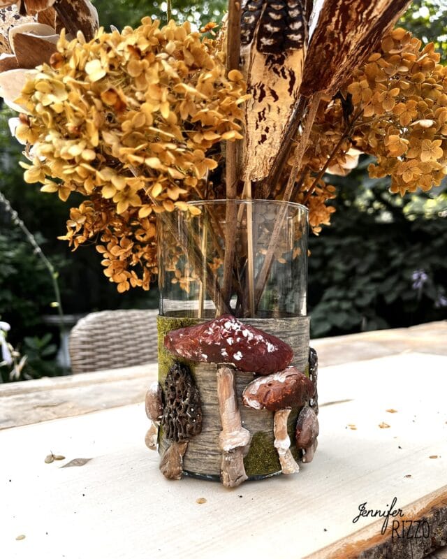 A clear vase decorated with faux mushrooms holds dried yellow flowers and tall dried plants on a wooden outdoor table.