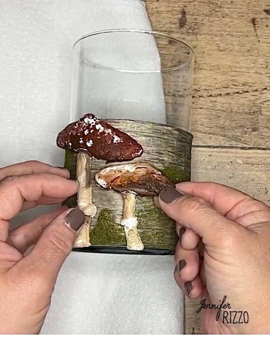 Hands attaching artificial mushrooms to a glass vase wrapped in faux bark and moss on a wooden surface.