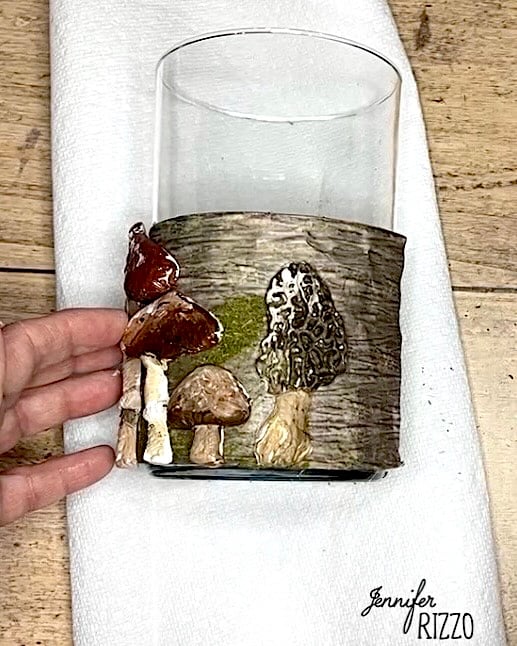A hand holds a glass candle holder decorated with clay mushrooms and birch bark texture. The name “Jennifer Rizzo” appears at the bottom right.