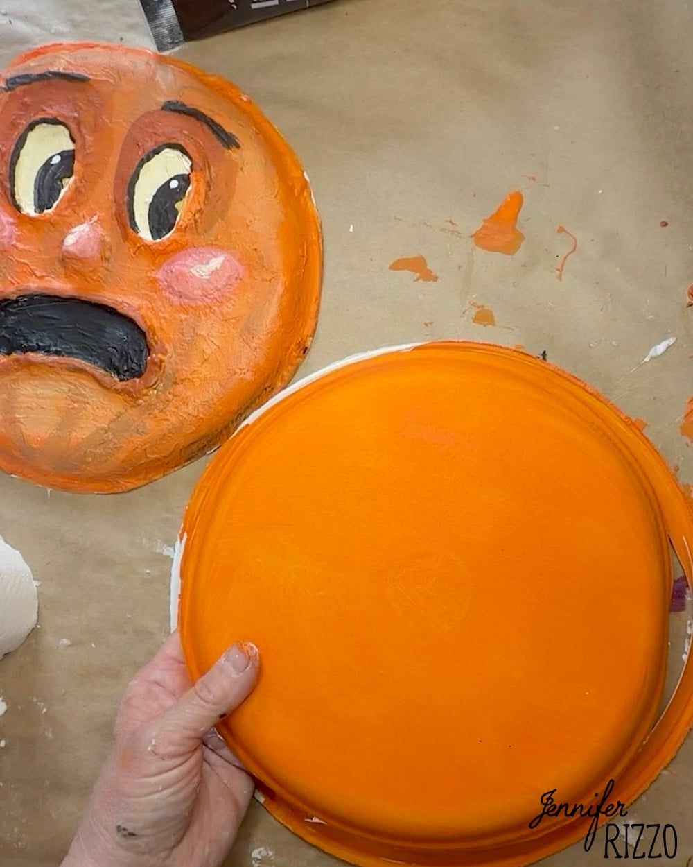 A hand holds a round object painted orange next to a painted face with wide eyes and an open mouth, on a paint-splattered surface.