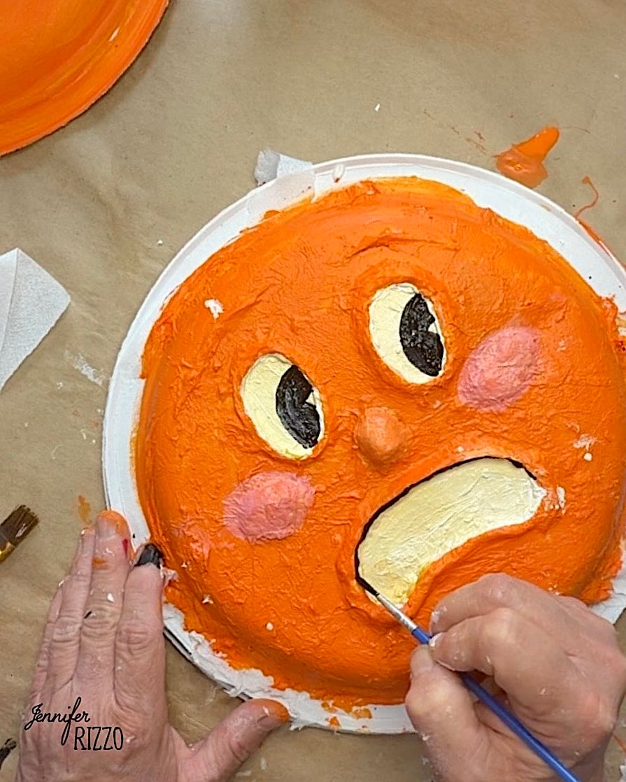 A person paints the mouth of an orange, round face with large eyes and pink cheeks on a paper plate using a small brush.