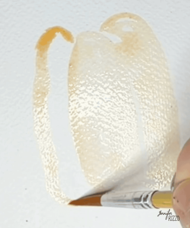 A hand uses a paintbrush to apply light orange watercolor paint in the shape of a pumpkin on textured white paper.