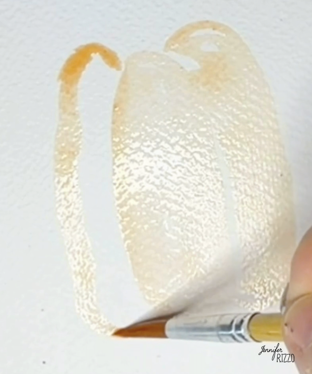 A hand uses a paintbrush to apply light orange watercolor paint in the shape of a pumpkin on textured white paper.