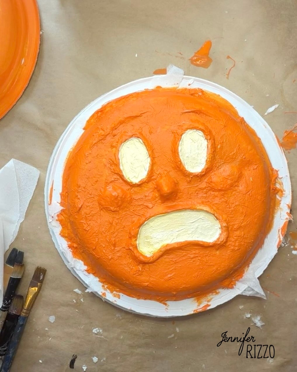 A round object painted orange with white eyes and a frown, resembling a distressed emoji face, sits on a paper plate next to paintbrushes and paper towels.