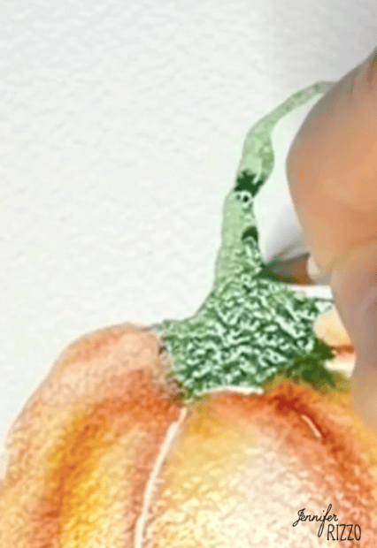 A hand uses a paintbrush to add green watercolor paint to the stem of an orange pumpkin illustration on textured paper.