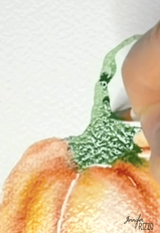 A hand uses a paintbrush to add green watercolor paint to the stem of an orange pumpkin illustration on textured paper.