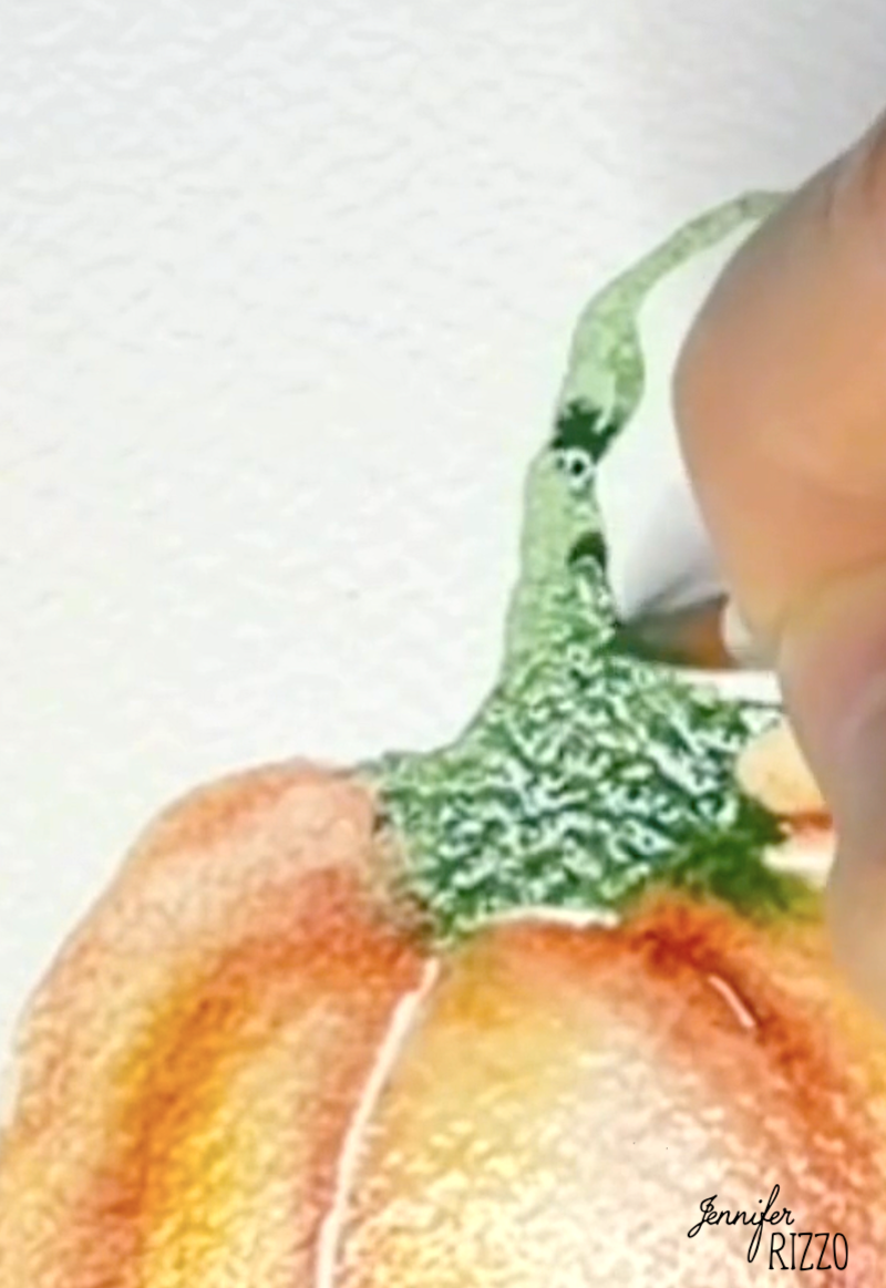 A hand uses a paintbrush to add green watercolor paint to the stem of an orange pumpkin illustration on textured paper.