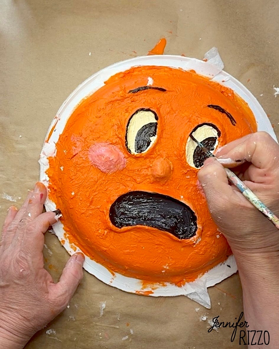 Person painting facial details on an orange cake designed to look like a surprised or scared emoji face.