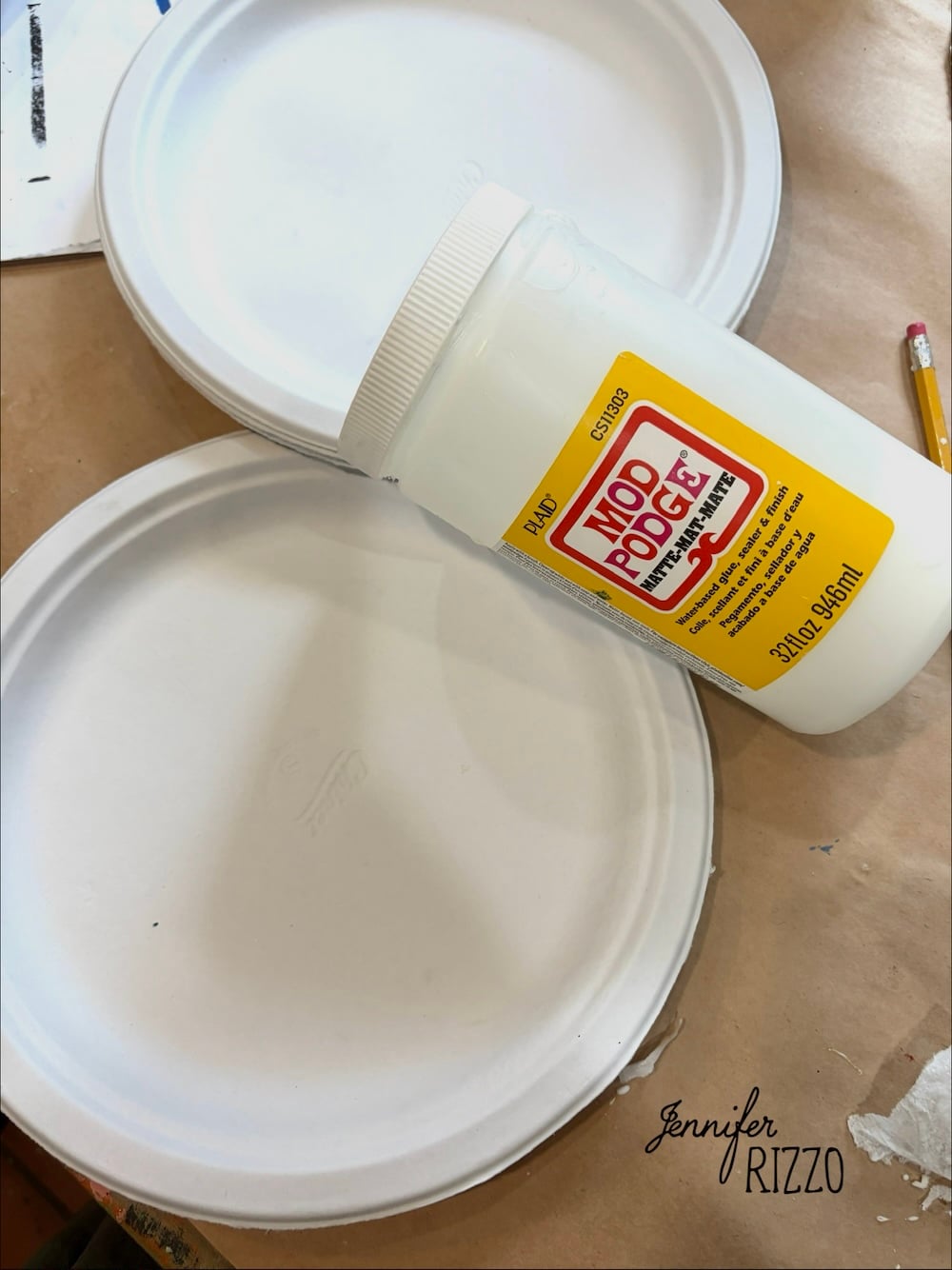 A container of Mod Podge rests on three stacked white paper plates next to a pencil, all placed on a brown surface.