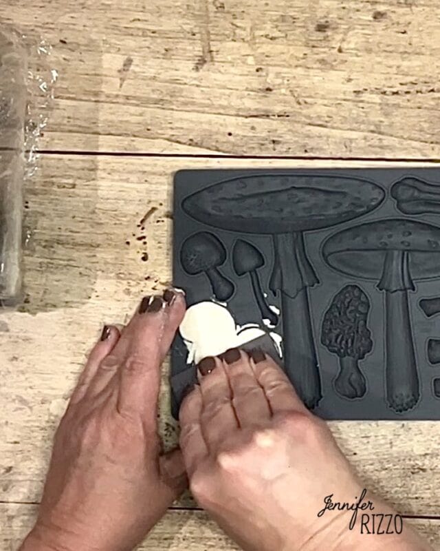 Two hands press white clay into a gray silicone mold with various mushroom shapes on a wooden surface.