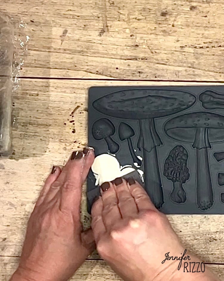 Two hands press white clay into a gray silicone mold with various mushroom shapes on a wooden surface.