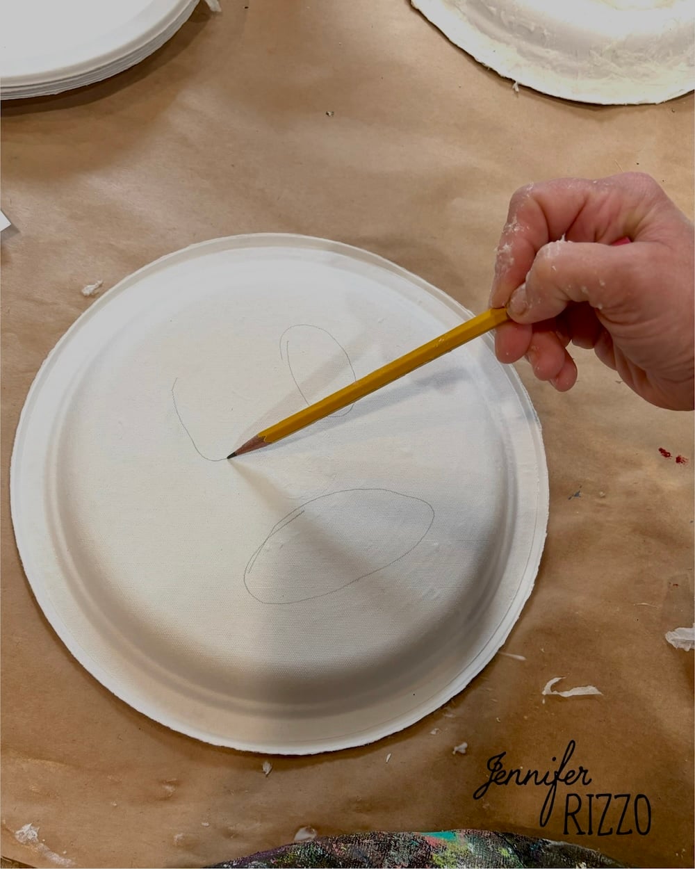 A hand holding a pencil is sketching shapes on a white paper plate placed on a brown surface. The name “Jennifer Rizzo” appears in the lower right corner.