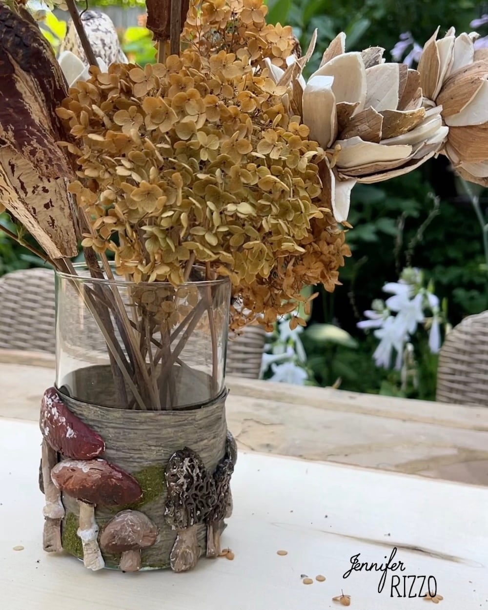 A glass vase with dried flowers sits on a table, decorated with small mushroom sculptures at its base. The background features outdoor greenery and wicker chairs.
