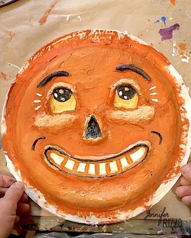 A person holds a textured, orange, round paper plate painted with a smiling face, featuring large eyes, arched brows, and visible teeth.