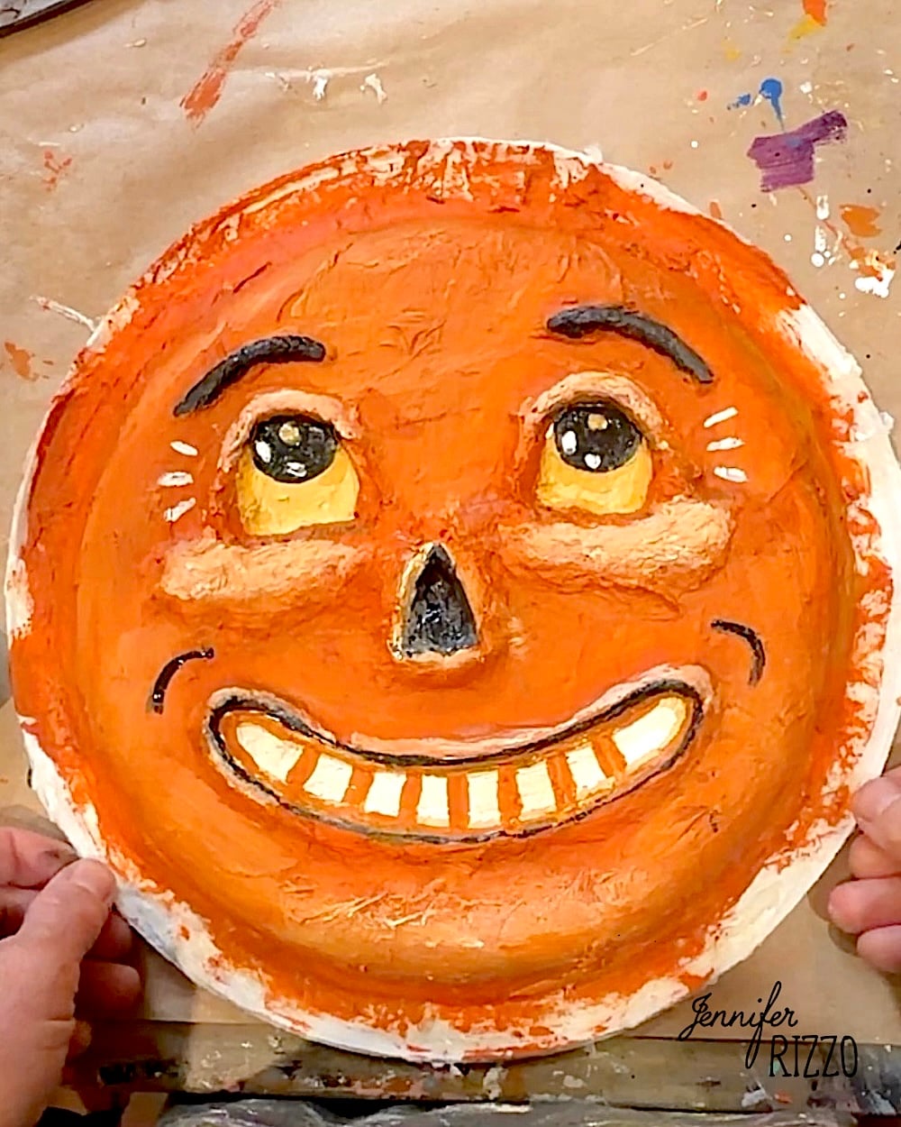 A person holds a textured, orange, round paper plate painted with a smiling face, featuring large eyes, arched brows, and visible teeth.