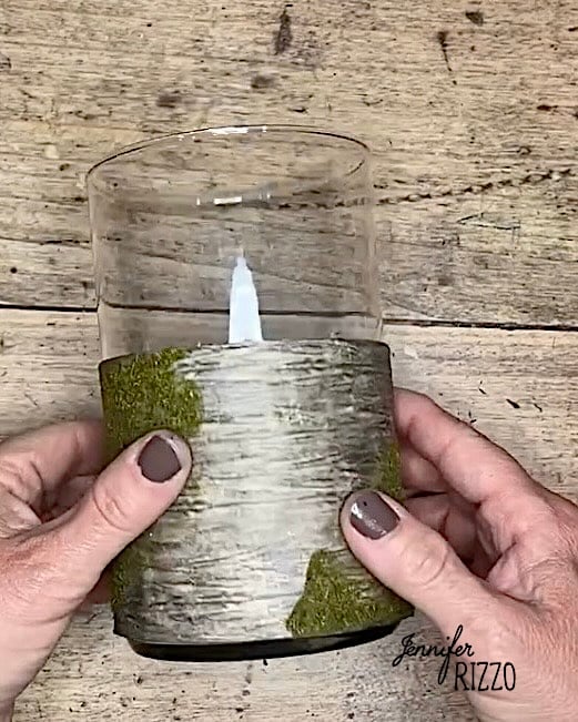 Two hands holding a glass candle holder decorated with birch bark and moss, placed on a wooden surface.