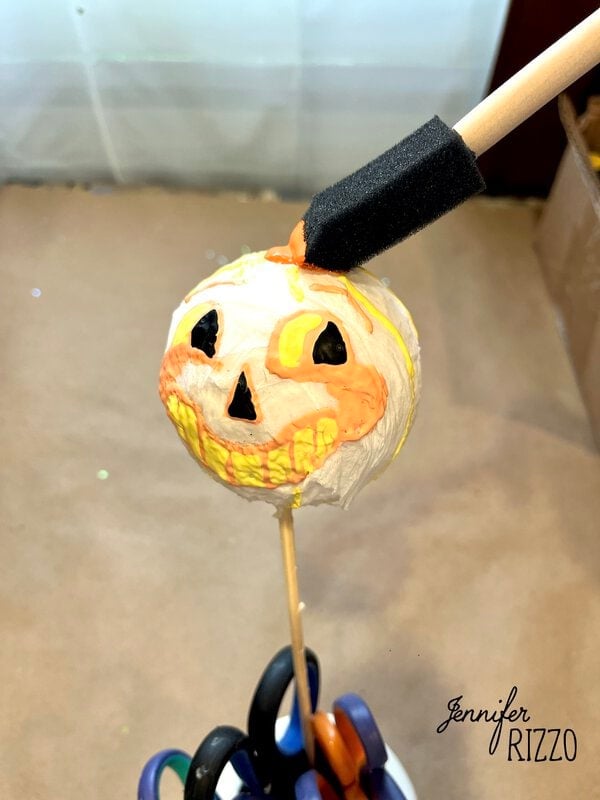 A foam brush paints a small pumpkin face on a round object mounted on a stick, with “Jennifer Rizzo” text in the corner.