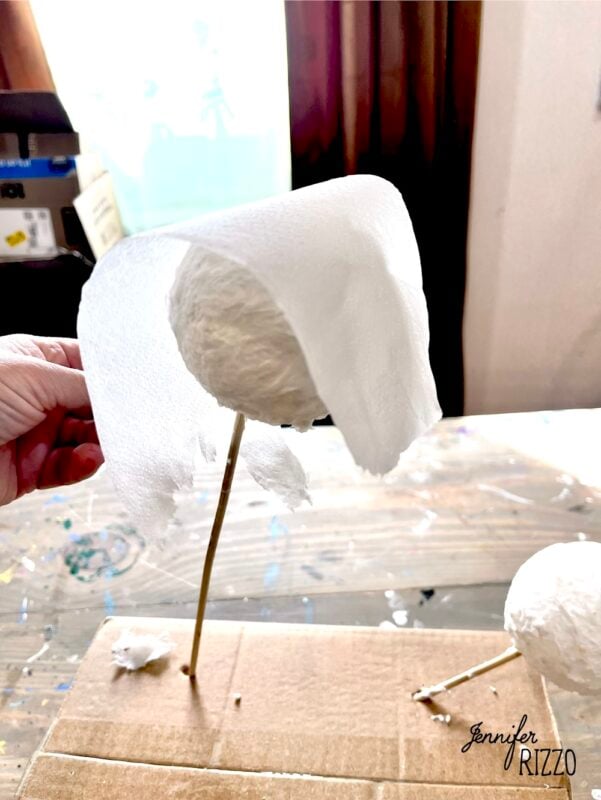 A hand places a white paper towel over a round paper mache form on a stick, which is inserted into cardboard.