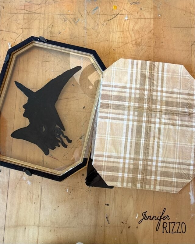 An octagonal frame with a glass panel featuring a black flying witch silhouette, next to a plaid-patterned backing, sits on a wooden surface. “Jennifer Rizzo” is printed in the corner.