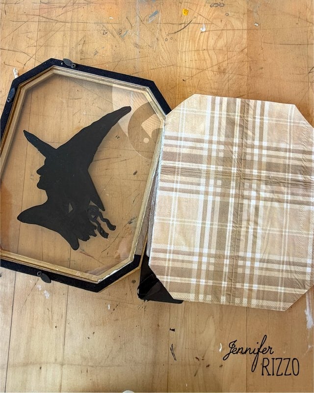 An octagonal frame with a glass panel featuring a black flying witch silhouette, next to a plaid-patterned backing, sits on a wooden surface. “Jennifer Rizzo” is printed in the corner.