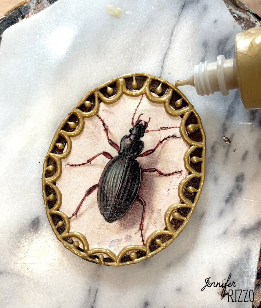 A hand applies glue to a gold oval frame holding an illustration of a black beetle on a marble surface.
