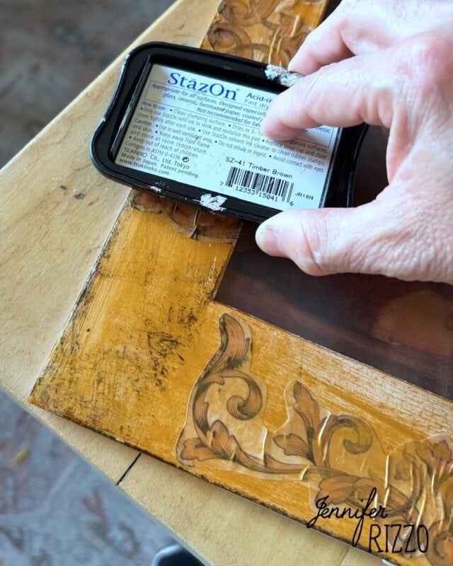 Hand holds a StazOn ink pad, applying brown ink to the edge of a wood picture frame with carved details.