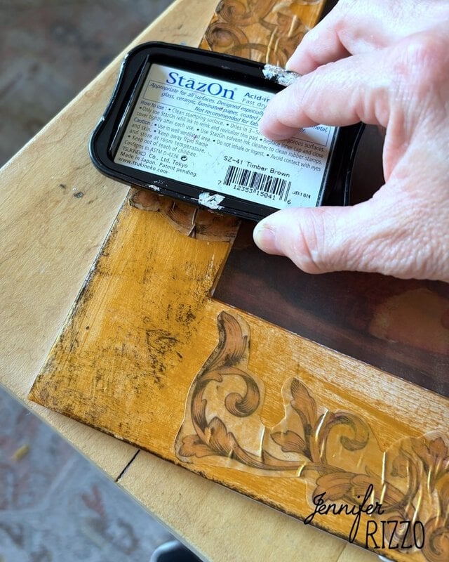 Hand holds a StazOn ink pad, applying brown ink to the edge of a wood picture frame with carved details.