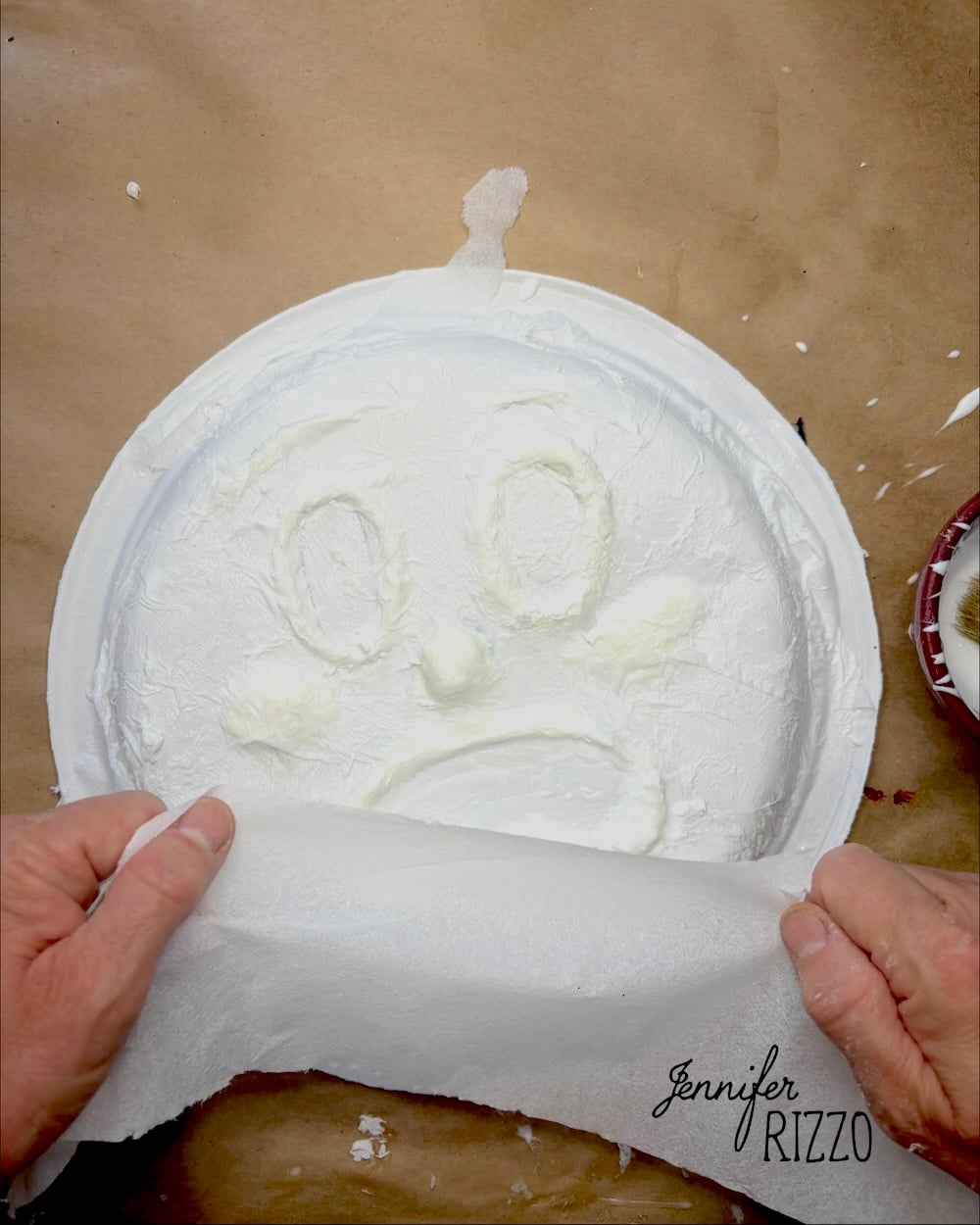 Hands peel paper from a white plate with a sad face drawn in a thick, textured substance. “Jennifer Rizzo” is written in the lower right corner.