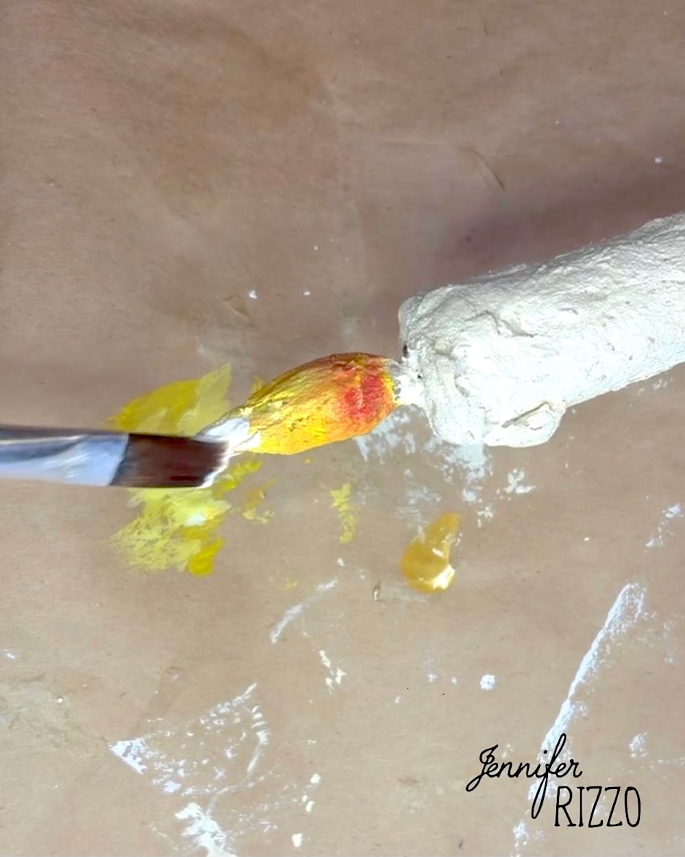 A paintbrush applies white paint to the yellow and red tip of a textured object on brown paper, with paint spots nearby.