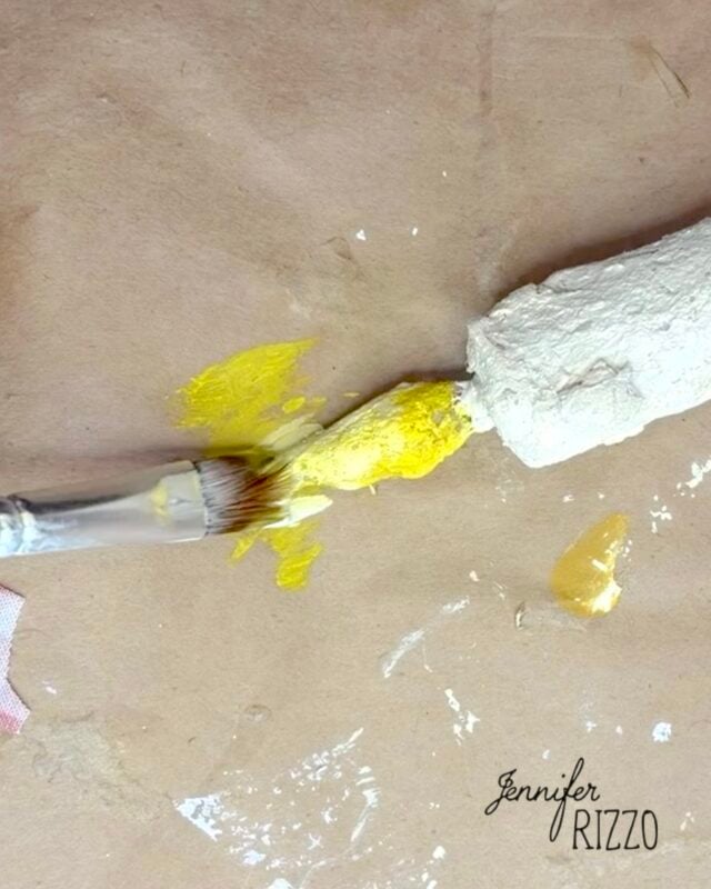 A paintbrush applies yellow paint to a textured white object on a brown paper surface. The name “Jennifer Rizzo” appears in the bottom right corner.