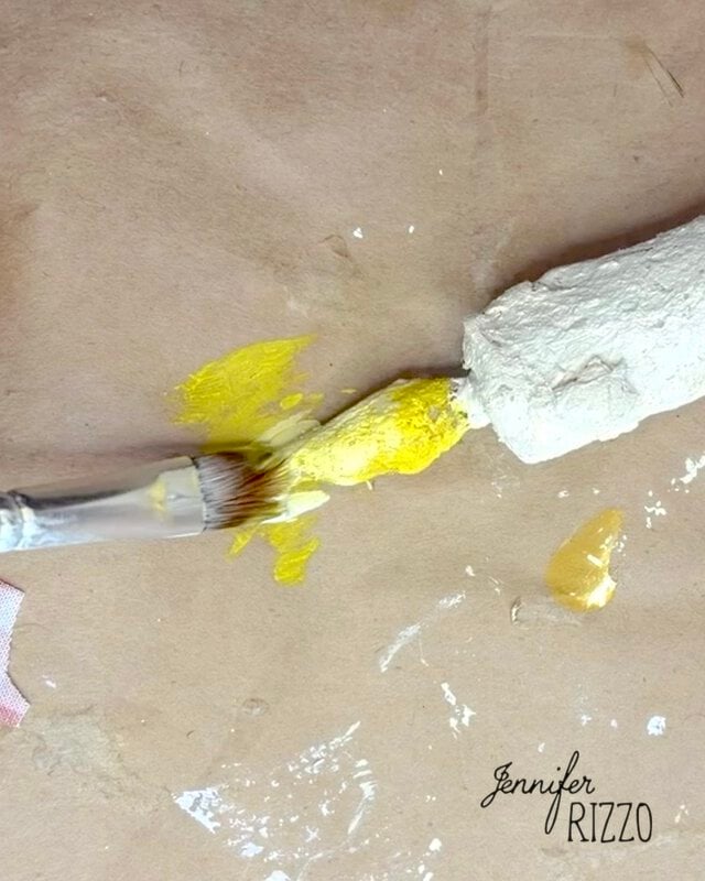 A paintbrush applies yellow paint to a textured white object on a brown paper surface. The name “Jennifer Rizzo” appears in the bottom right corner.