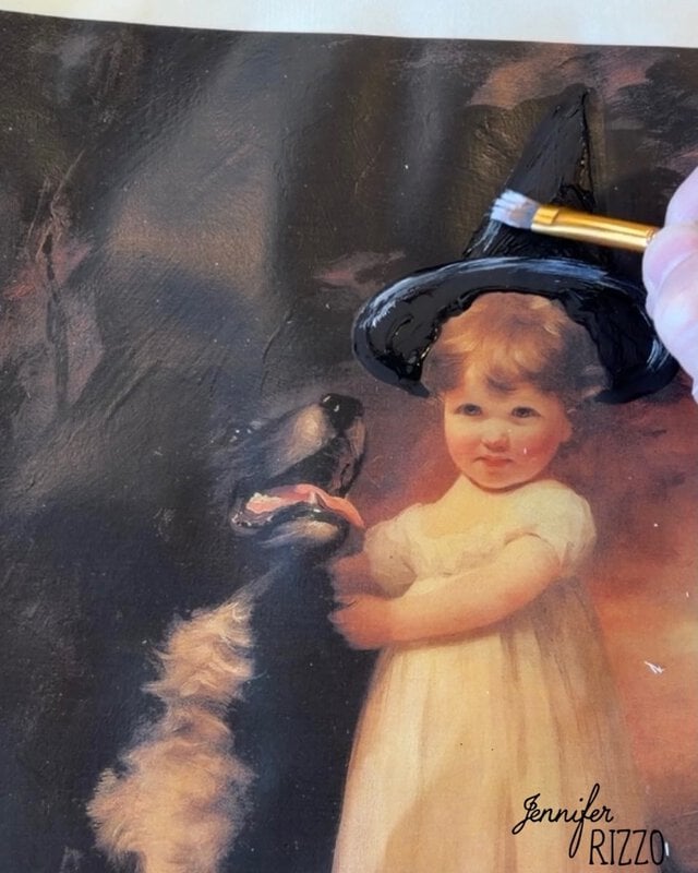 A hand paints a black witch hat on a portrait of a young child standing beside a dog. The painting is signed “Jennifer Rizzo” in the bottom right corner.