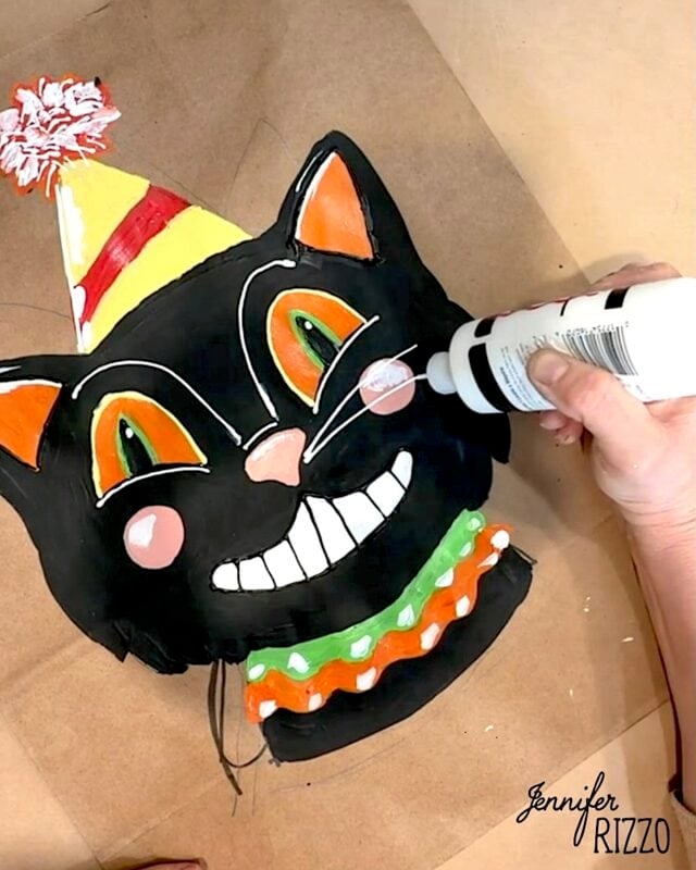 Person adds white paint to a colorful, smiling black cat face wearing a party hat, crafted on brown paper. The image features the signature “Jennifer Rizzo” in the lower right corner.