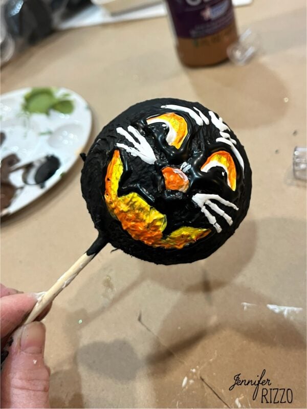 A hand holds a round object painted with a black cat face on a yellow-orange background, mounted on a stick. Art supplies are visible in the background.