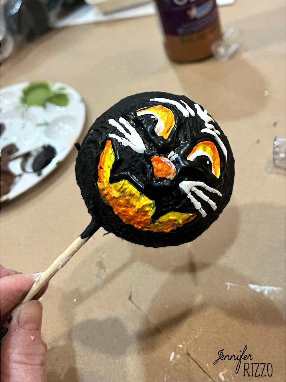A hand holds a round object painted with a black cat face on a yellow-orange background, mounted on a stick. Art supplies are visible in the background.