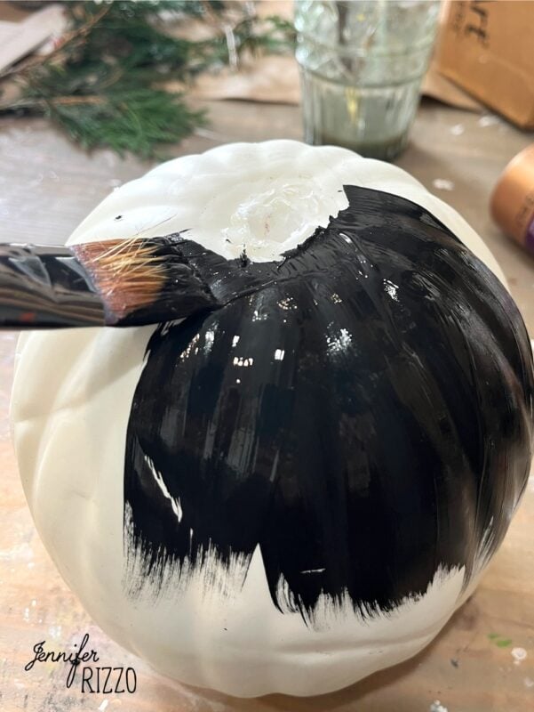 A paintbrush applies black paint to the surface of a white pumpkin. A glass jar and greenery are visible in the background.