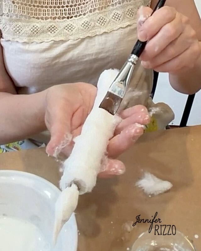 Person painting a fabric-wrapped object with white liquid using a brush, sitting at a table with crafting materials.