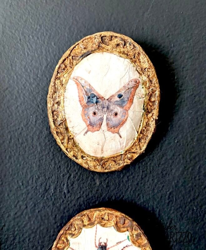A circular ornament with a painted butterfly is set in a textured gold frame against a black background.