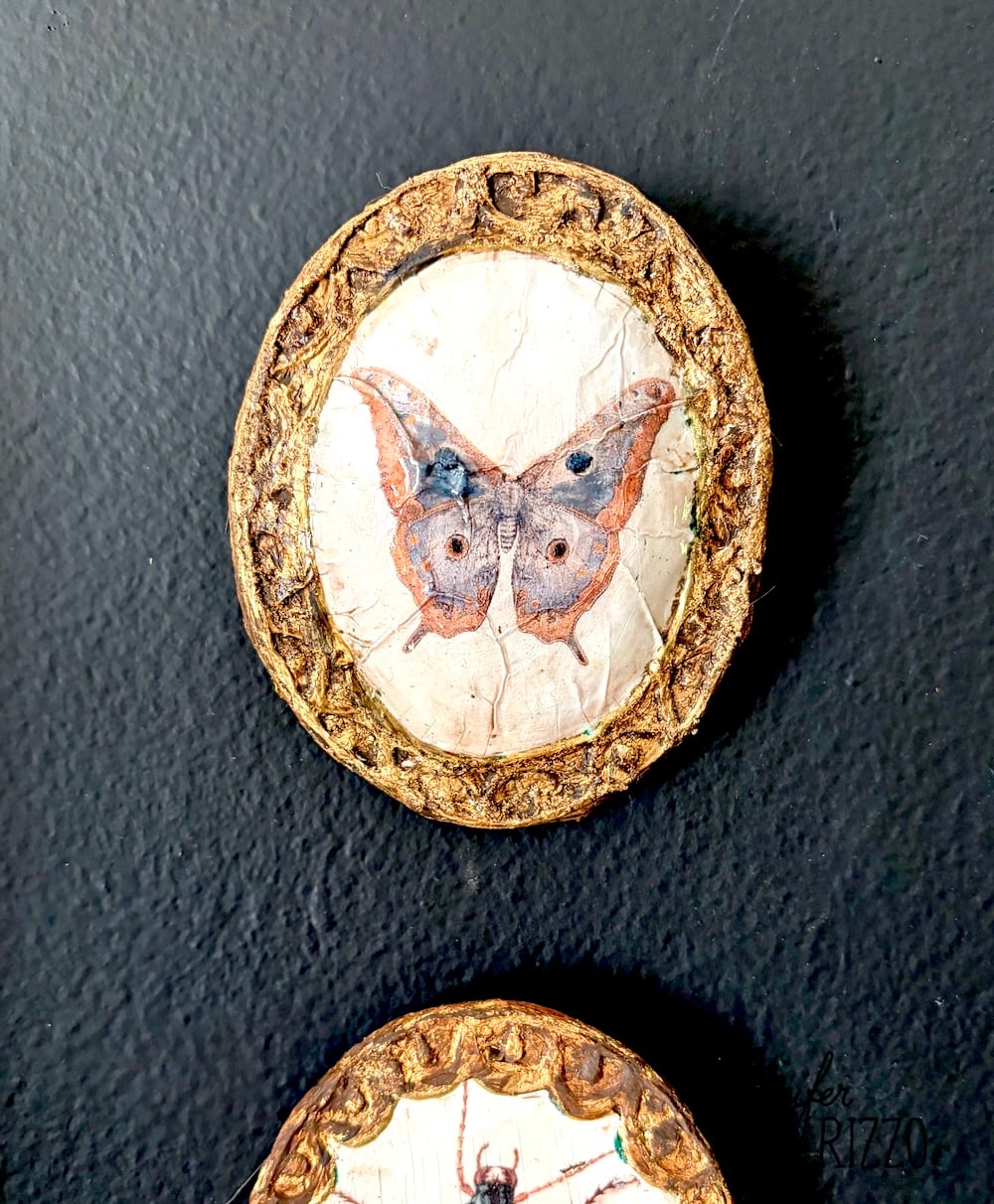 A circular ornament with a painted butterfly is set in a textured gold frame against a black background.