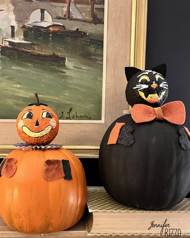Two Halloween-themed pumpkin decorations, one painted as a jack-o’-lantern with a collar, and the other as a black cat with a bow tie, displayed in front of a framed painting.