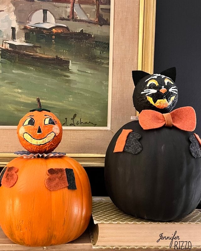 Two Halloween-themed pumpkin decorations, one painted as a jack-o’-lantern with a collar, and the other as a black cat with a bow tie, displayed in front of a framed painting.