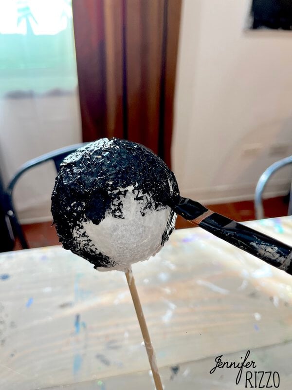 A brush paints black paint onto a white foam ball held on a wooden stick over a table, with chairs and curtains visible in the background.