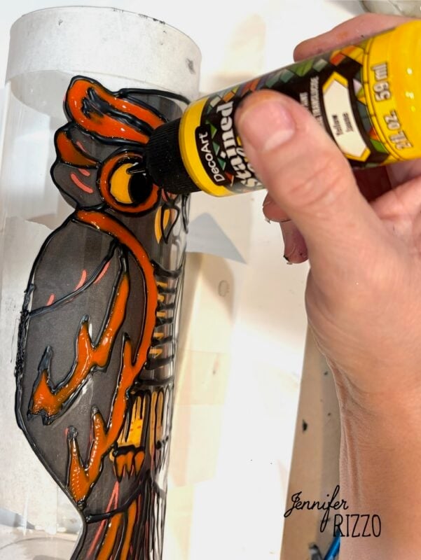 A hand uses yellow paint to color the eye of an owl design on a clear cylindrical surface.