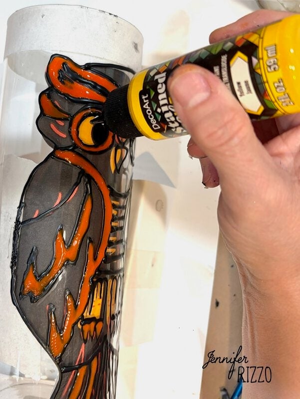 A hand uses yellow paint to color the eye of an owl design on a clear cylindrical surface.