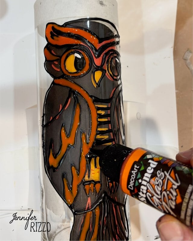 A hand applies orange glass paint to a black-outlined owl design on a vertical glass surface. The name Jennifer Rizzo appears in the bottom left corner.
