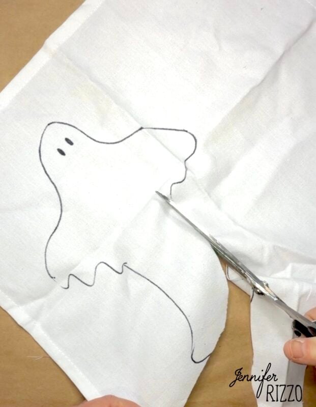 A hand uses scissors to cut out a ghost shape sketched on a piece of white fabric laying on a flat surface.