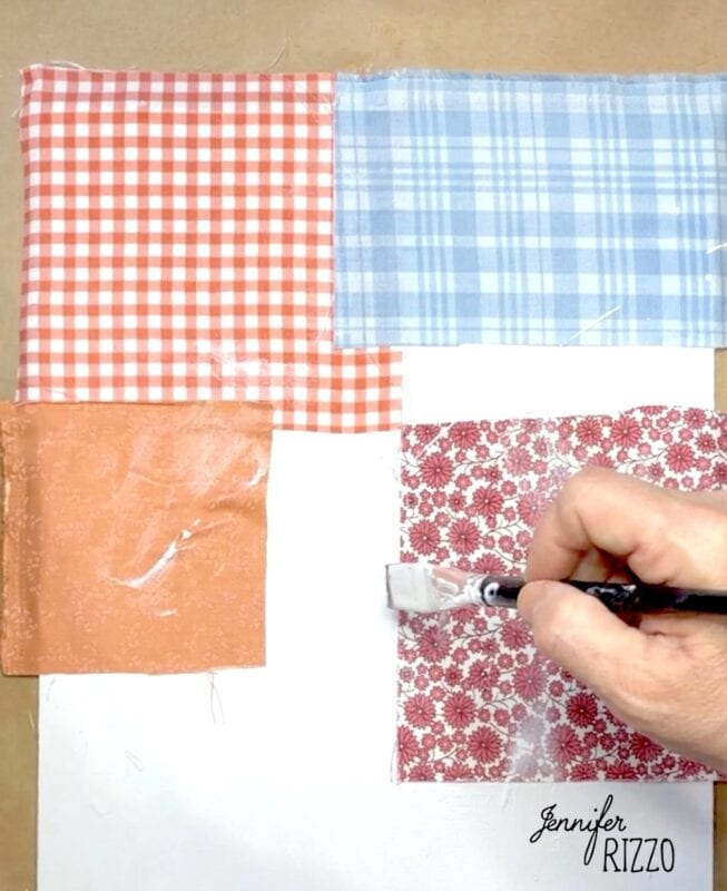 A hand uses a brush to apply glue or decoupage over four colorful fabric squares arranged on a white surface.
