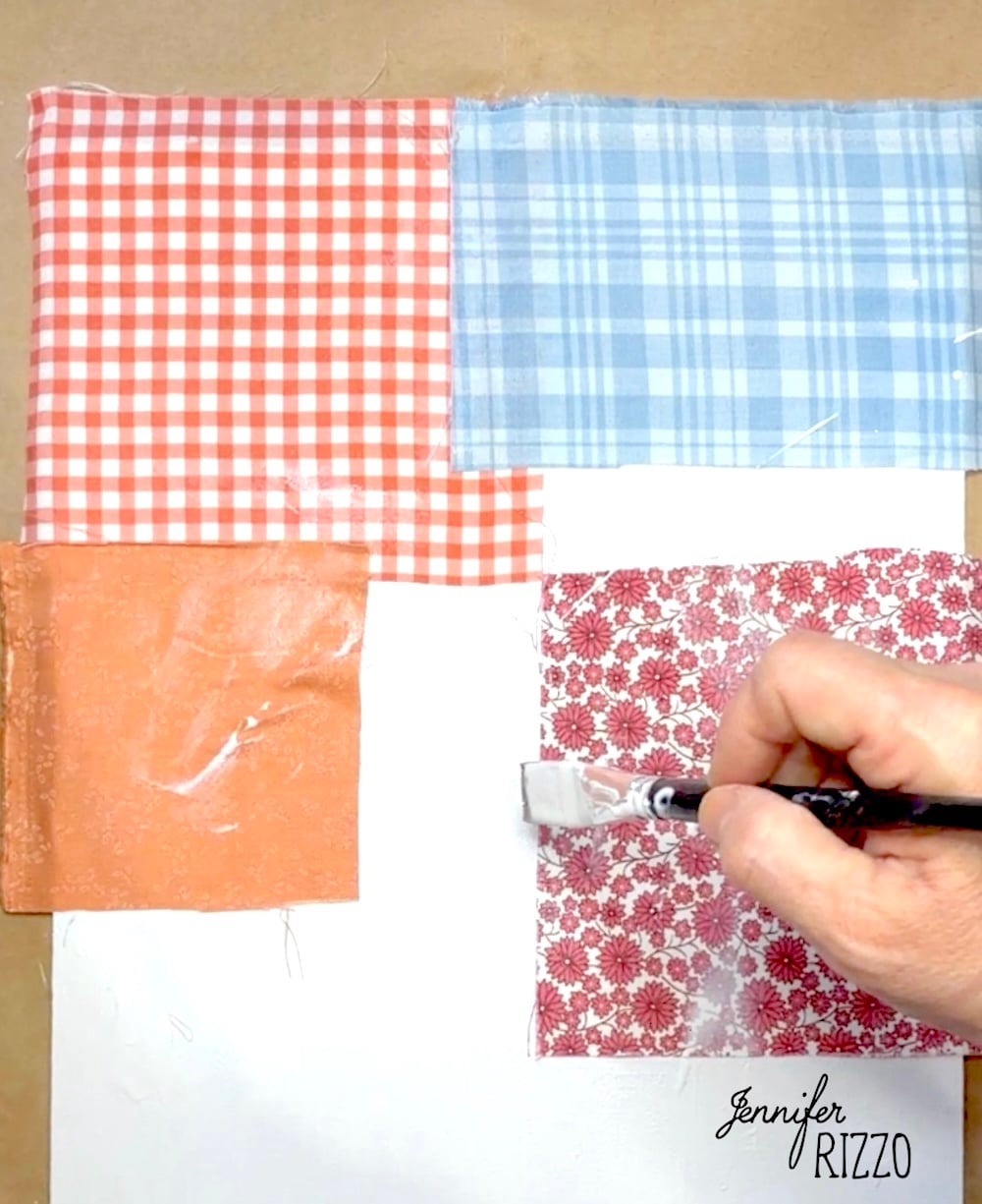 A hand uses a brush to apply glue or decoupage over four colorful fabric squares arranged on a white surface.