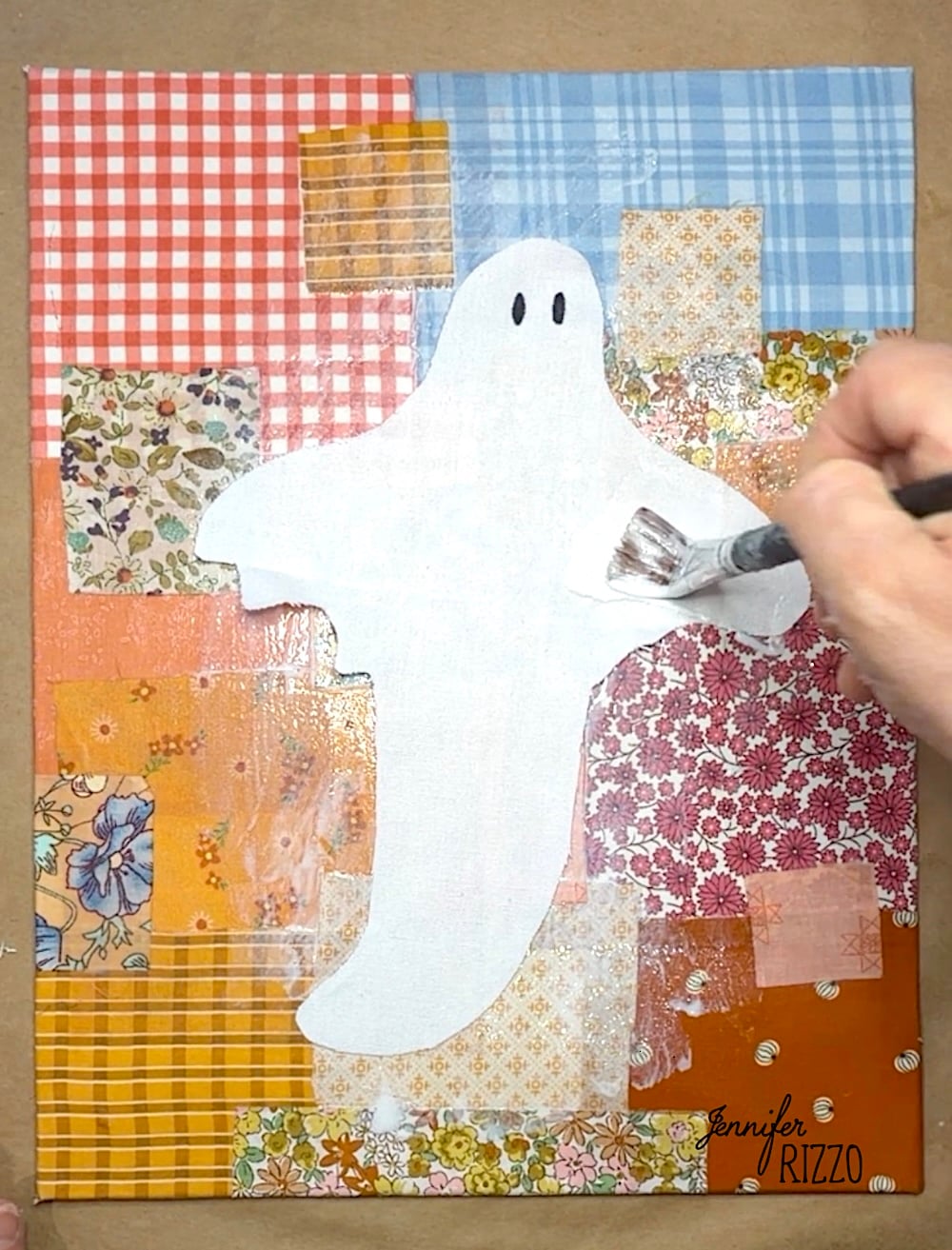 A hand paints a white ghost on a patchwork background of various patterned and colored squares on a canvas.