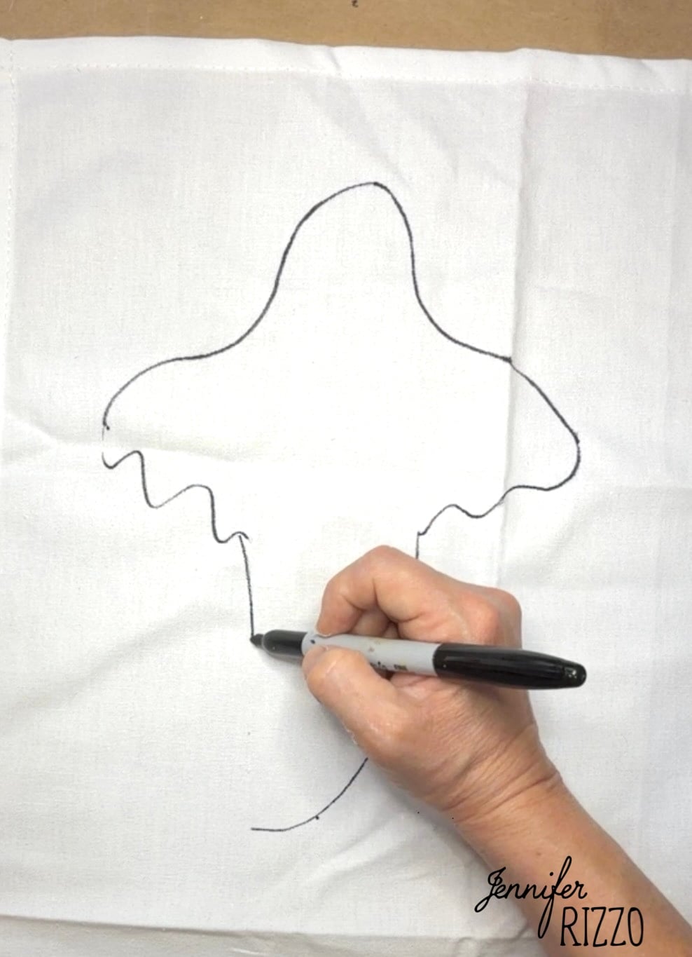 A hand draws the outline of a mushroom on white fabric with a black marker. The name “Jennifer Rizzo” appears in the bottom right corner.